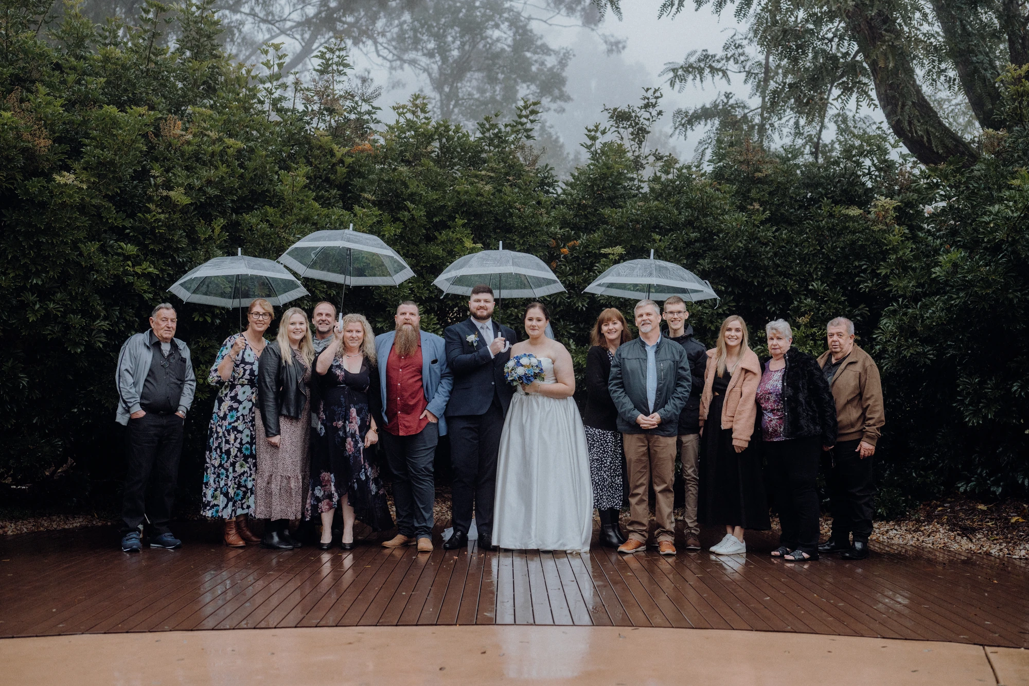 Newlyweds pose with family members beneath clear umbrellas.
