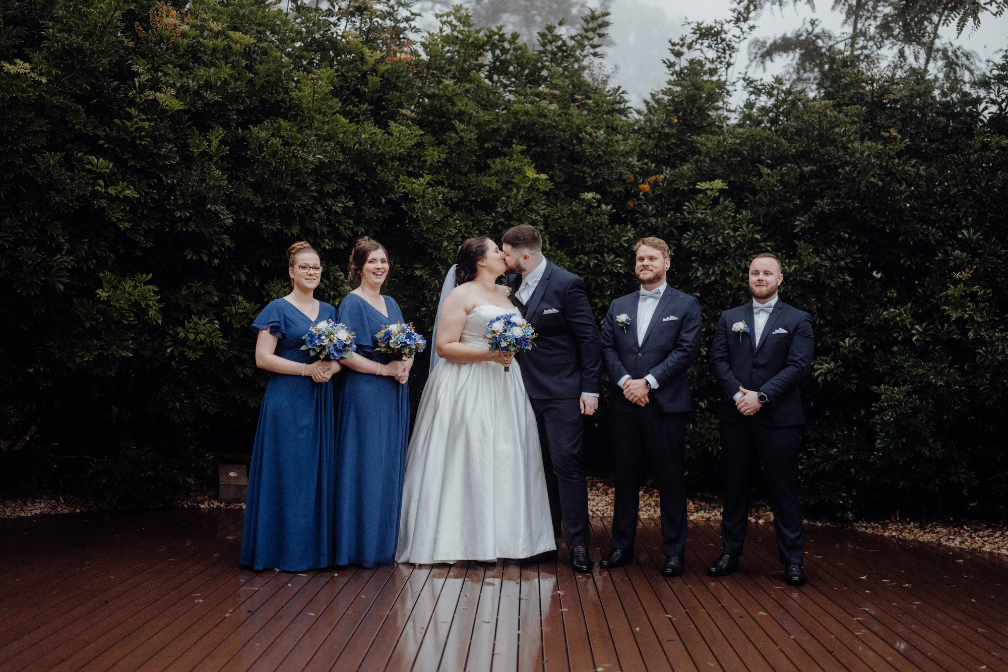 A couple kiss while their wedding party stands beside them.