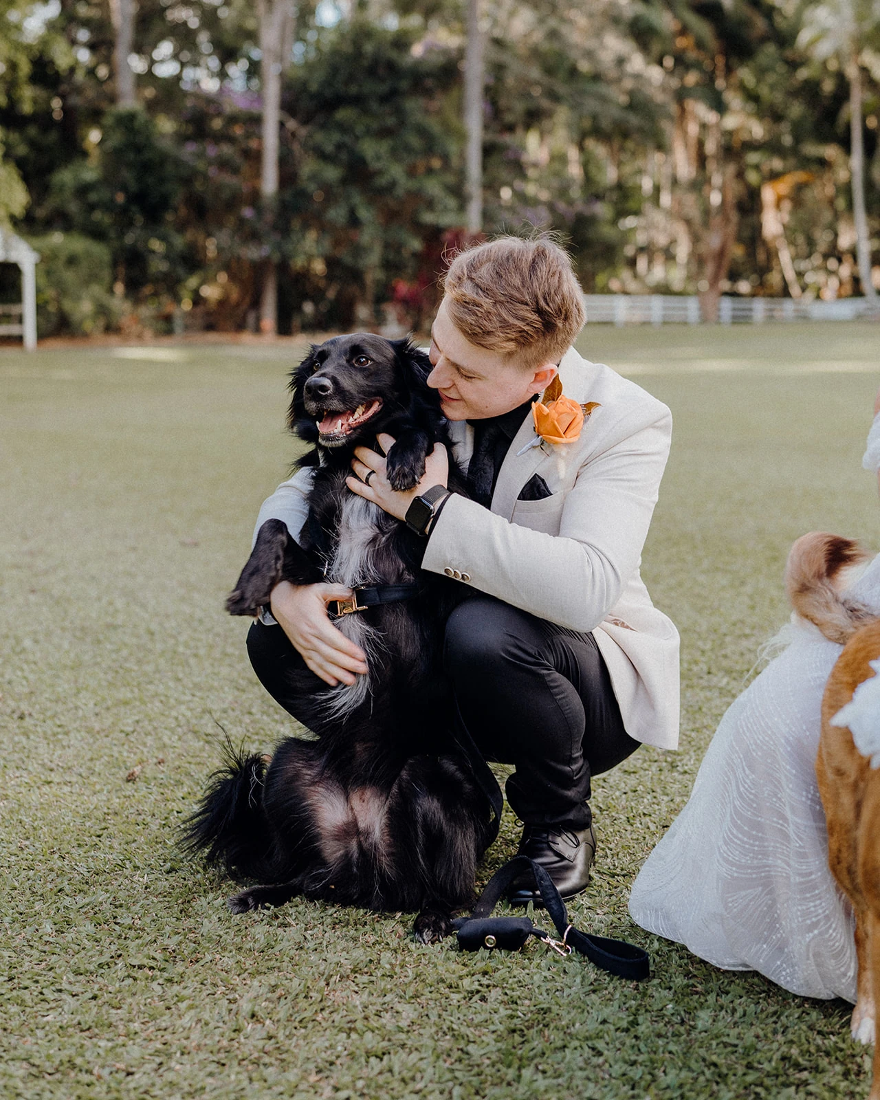 A groom kneels to cuddle a black dog on the lawn.