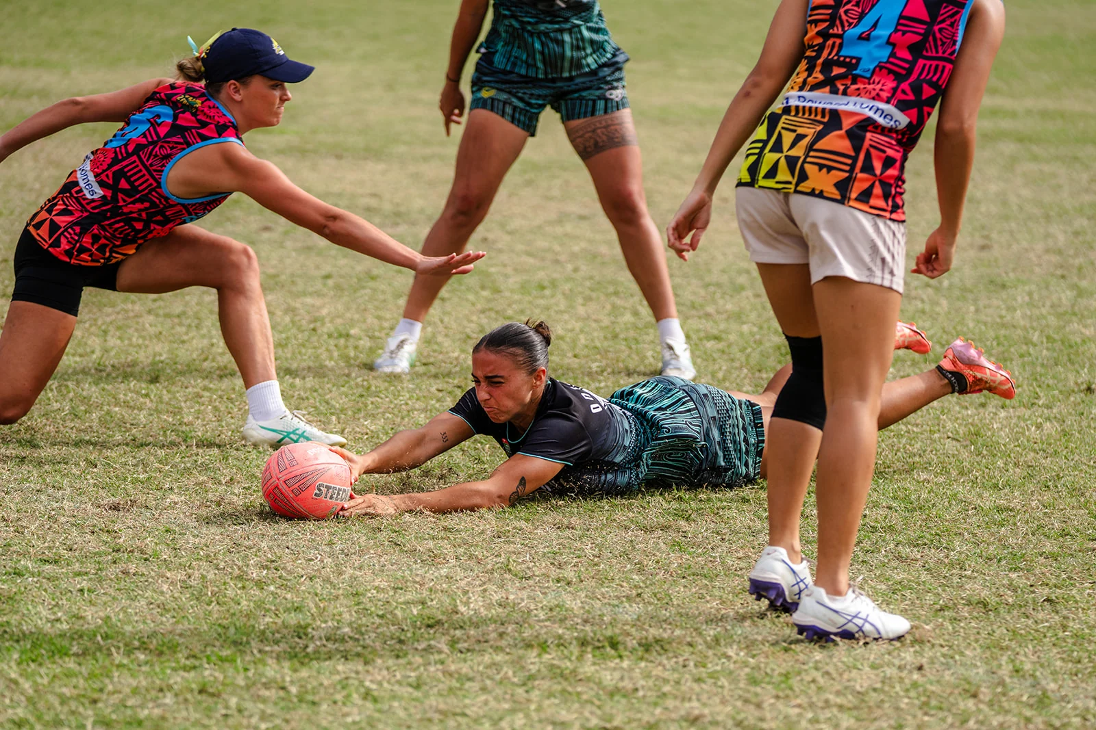 A woman stretches across the turf toward a pink ball as a defender reaches down.