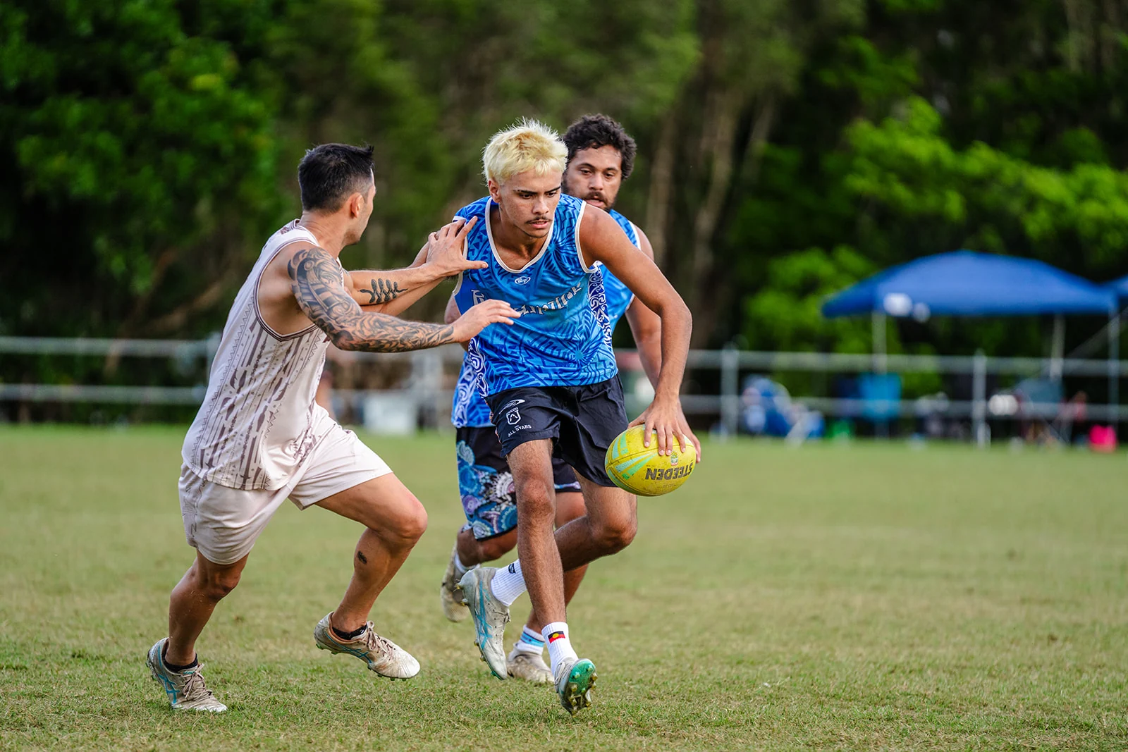 A player in blue carries the ball through two reaching defenders.