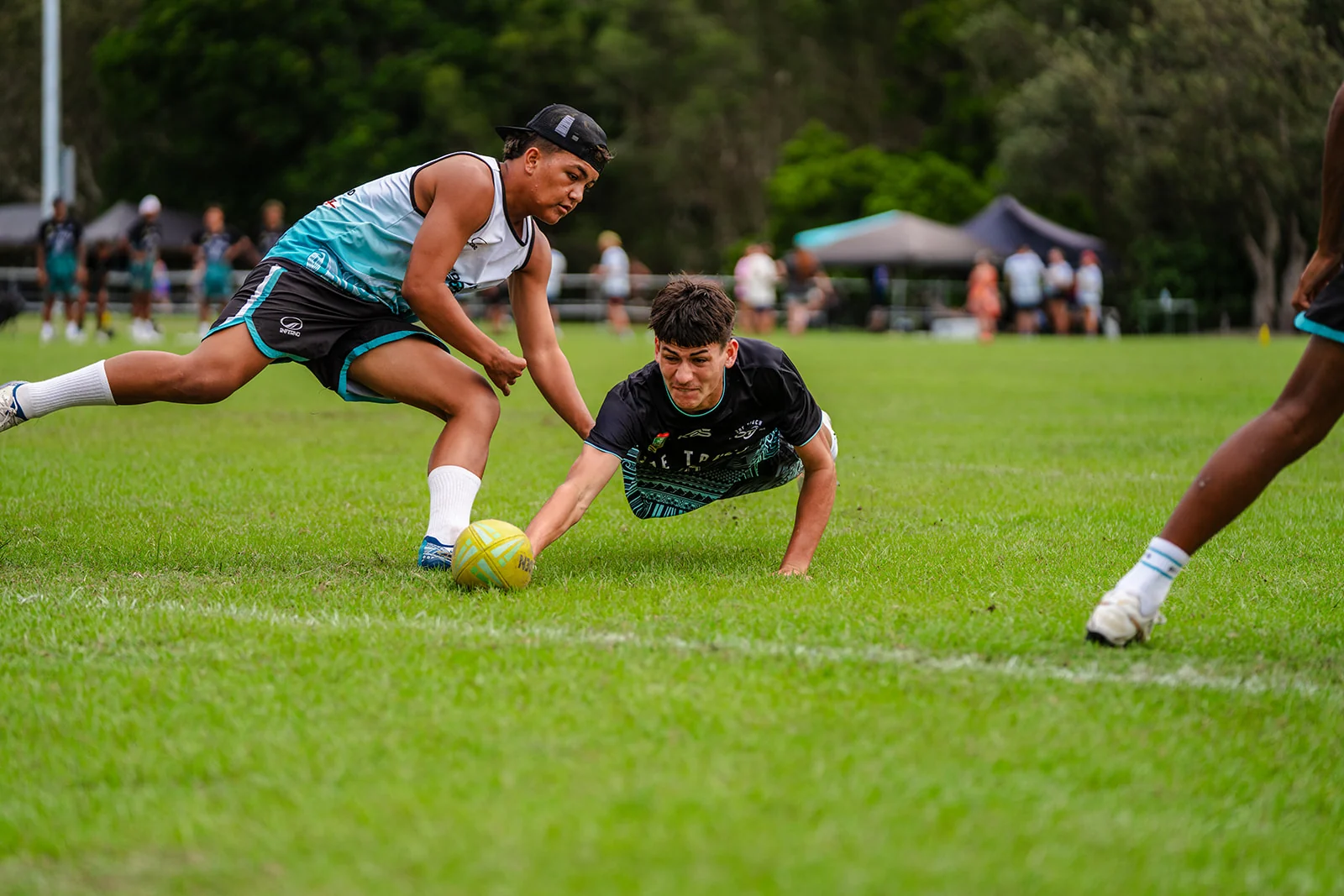 A diving ball carrier places the ball on the turf beneath a reaching opponent.