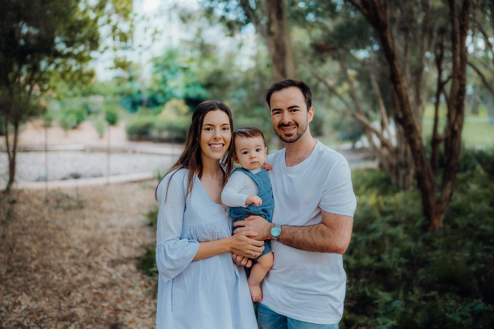 Parents hold their baby on a shaded woodland path.