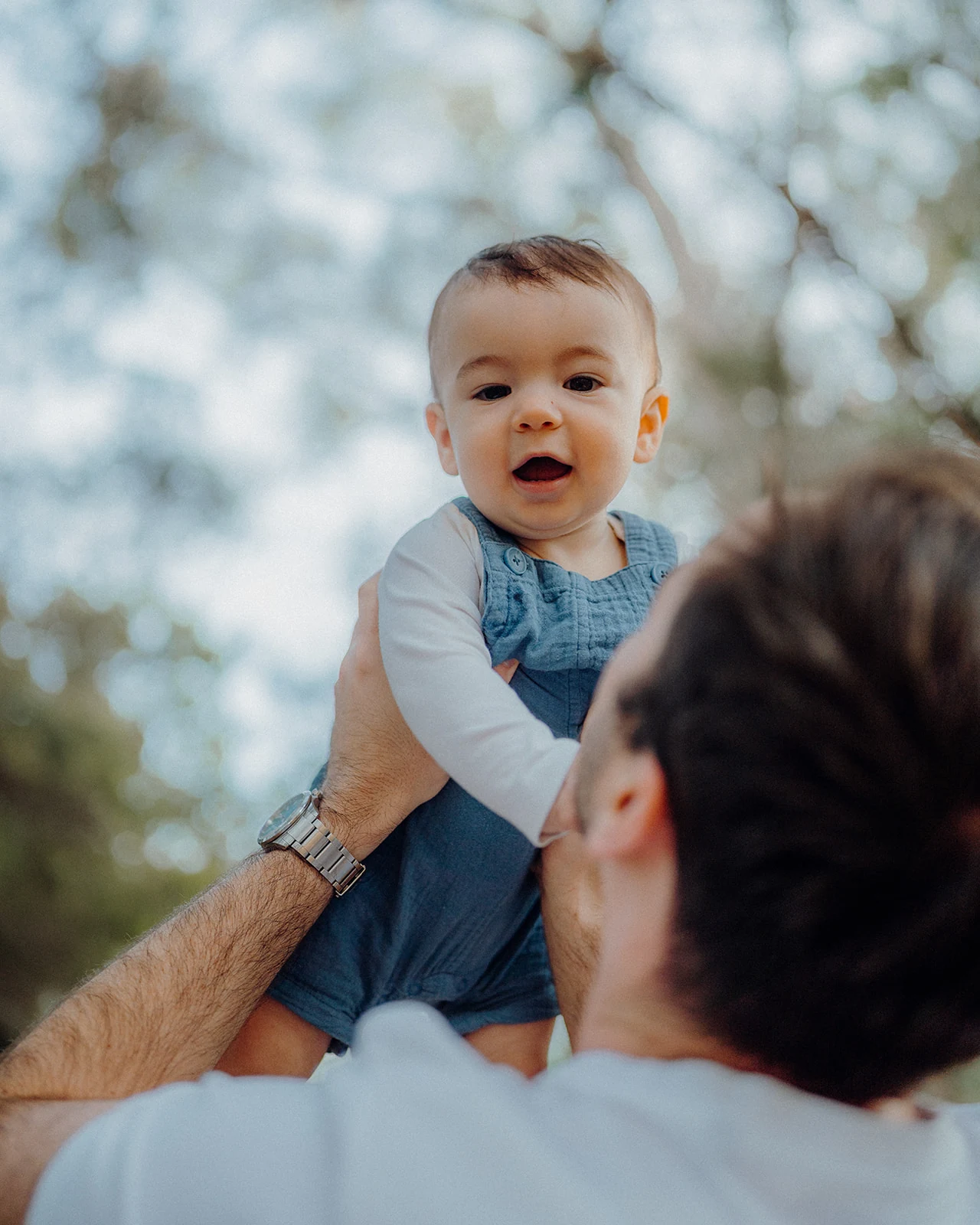 A father lifts his smiling baby beneath the trees.