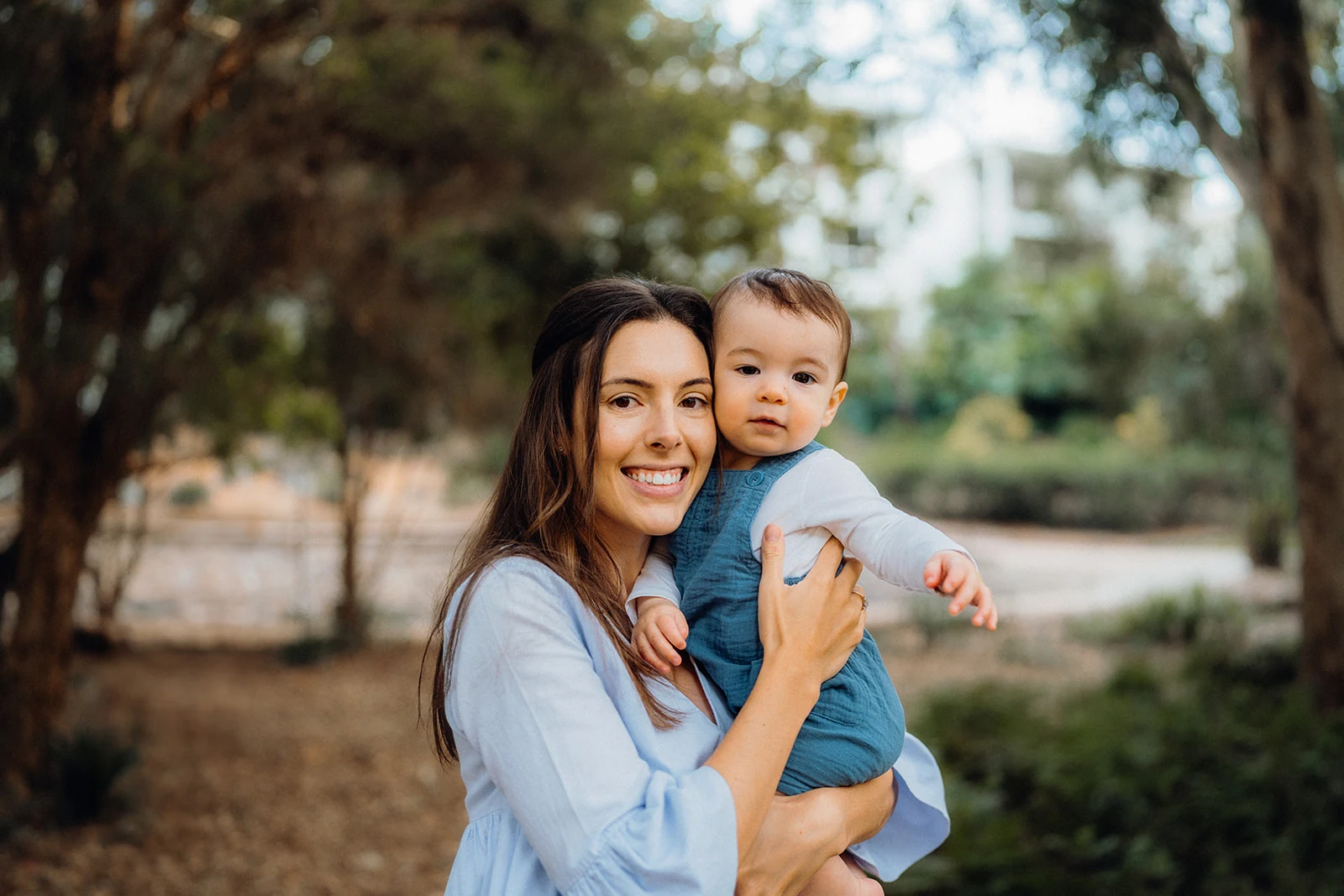 A mother holds her baby on a tree-lined path.