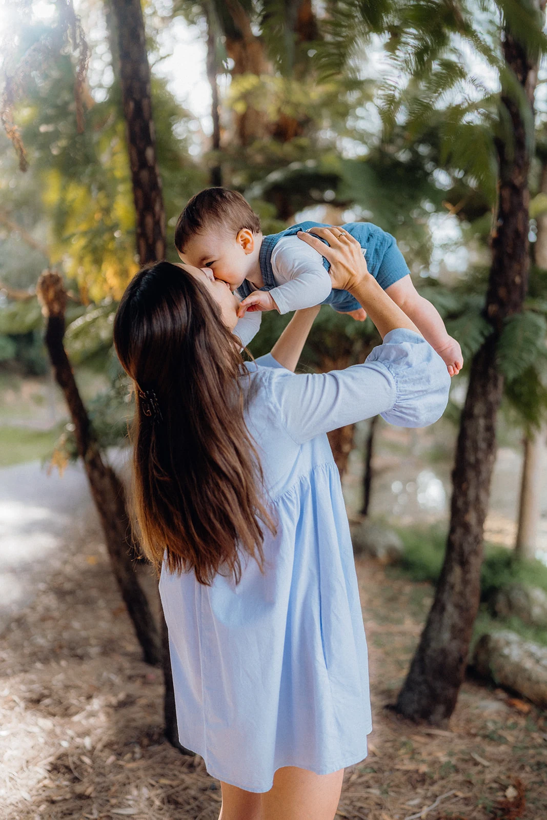 A mother lifts her baby for a kiss beneath the trees.