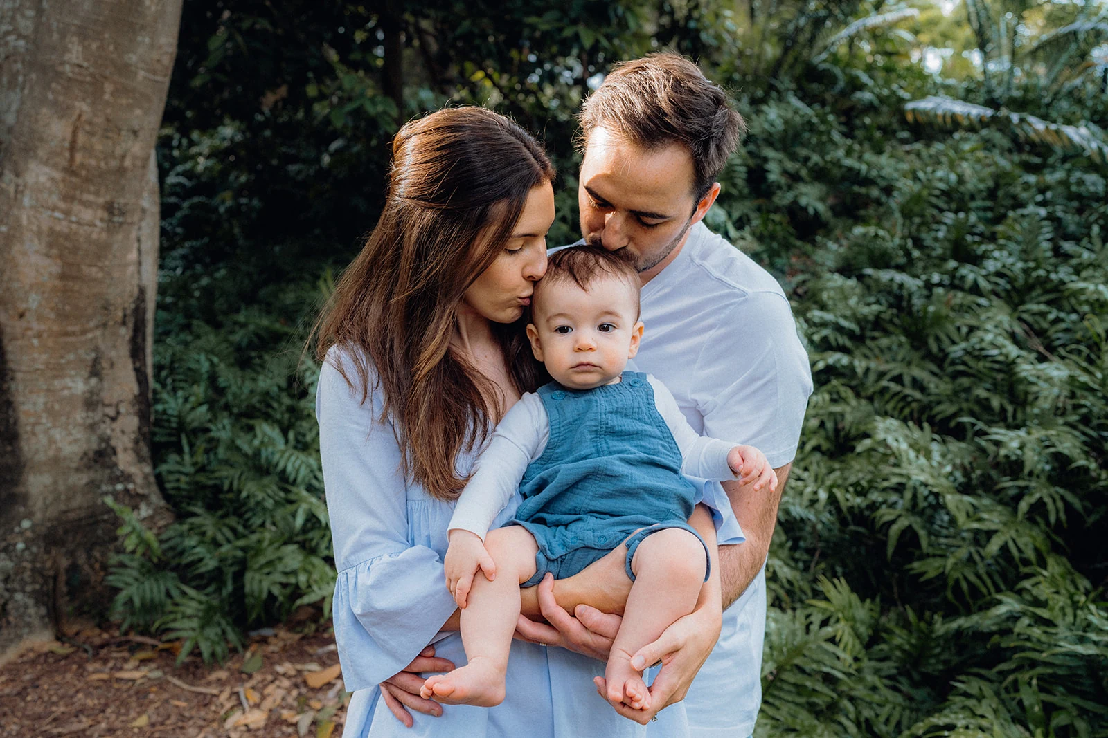 Parents kiss their baby’s head among green foliage.