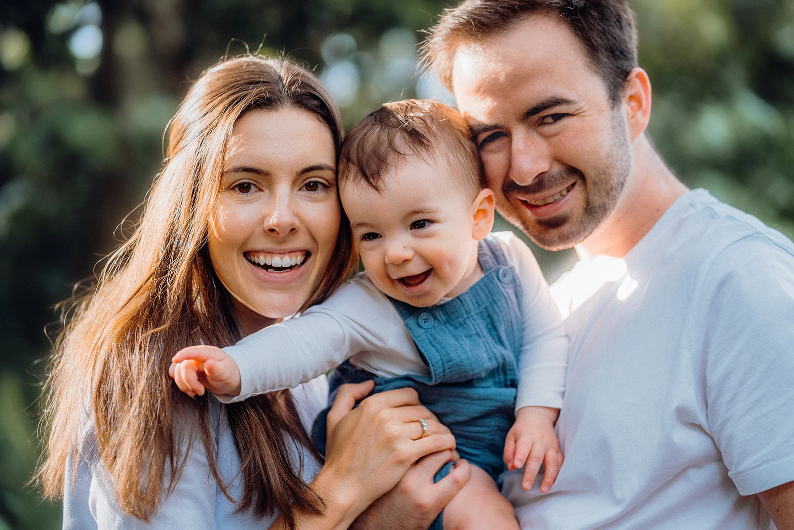 Parents and their laughing baby lean close together in warm sunlight.