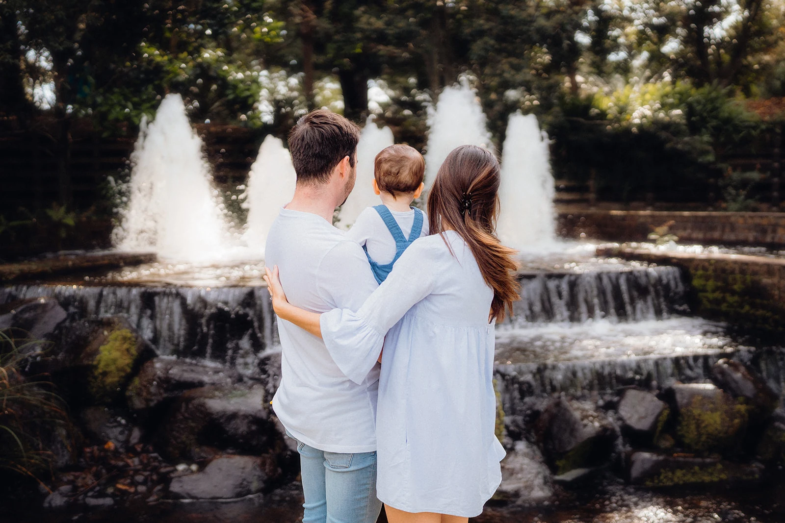 Parents hold their baby while watching a garden fountain.