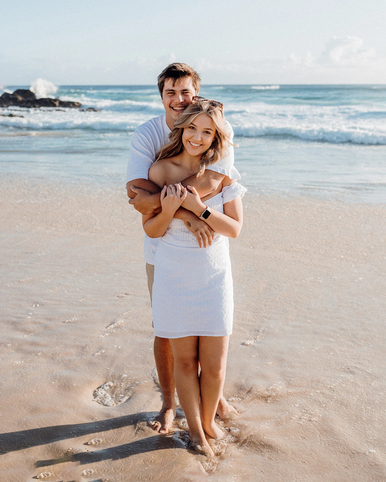 A couple embraces on the beach with the surf behind them.