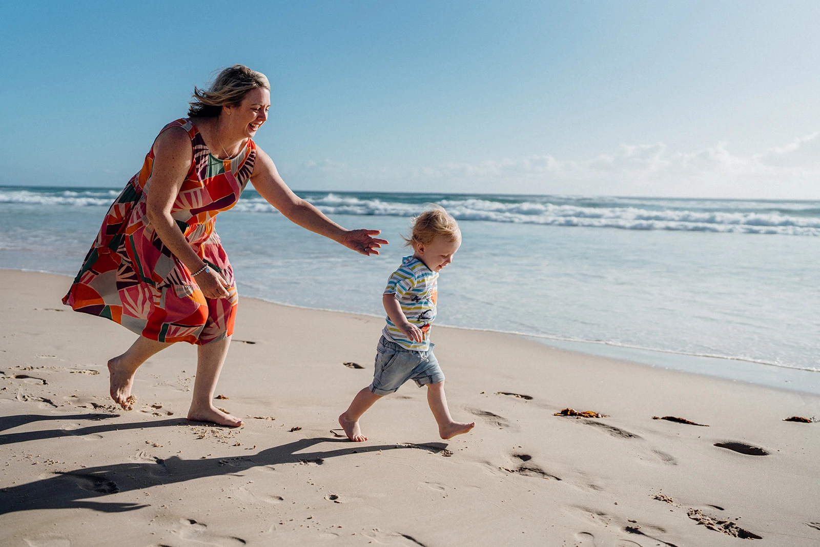 A woman runs beside a toddler on the sand.