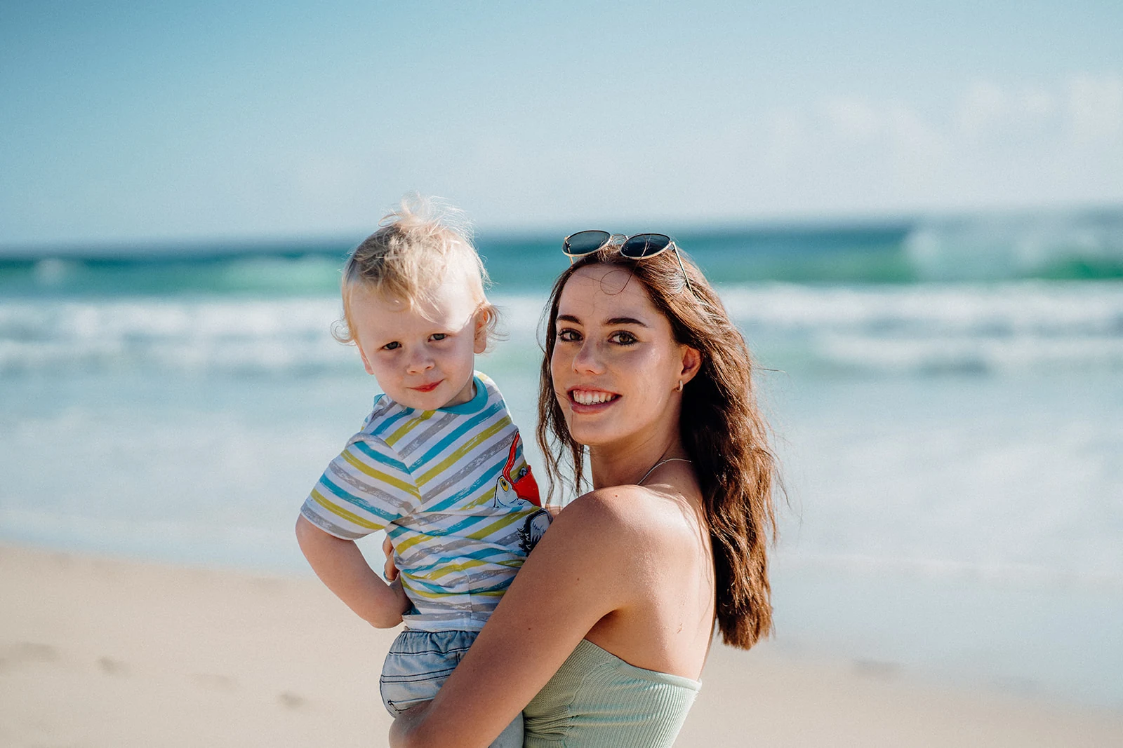 A woman holds a young child in front of turquoise surf.