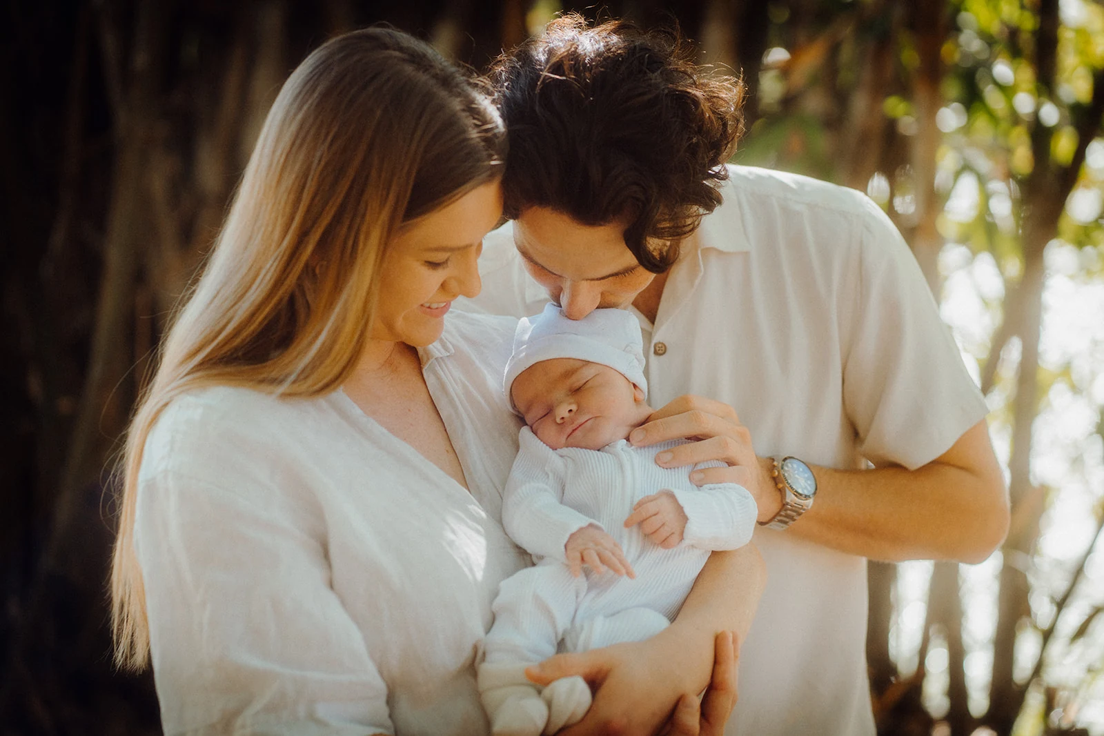 Parents lean close to their sleeping newborn beneath the trees.