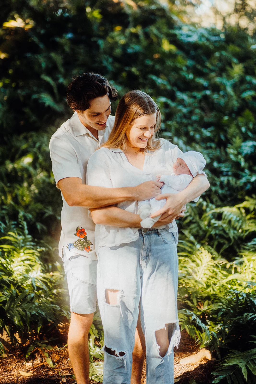 Parents hold their newborn together in a sunlit garden.