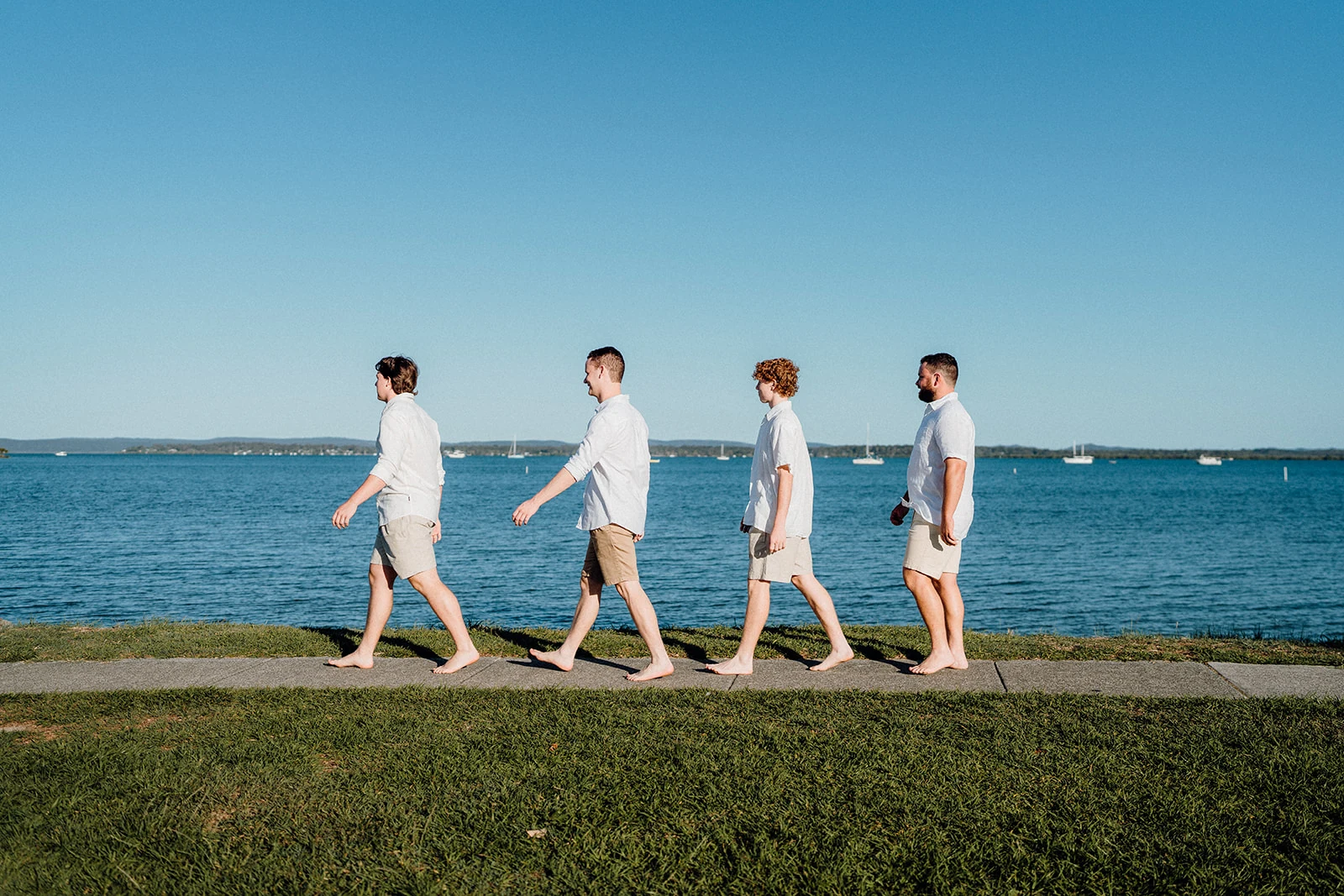 Four brothers walk in line beside the blue water.