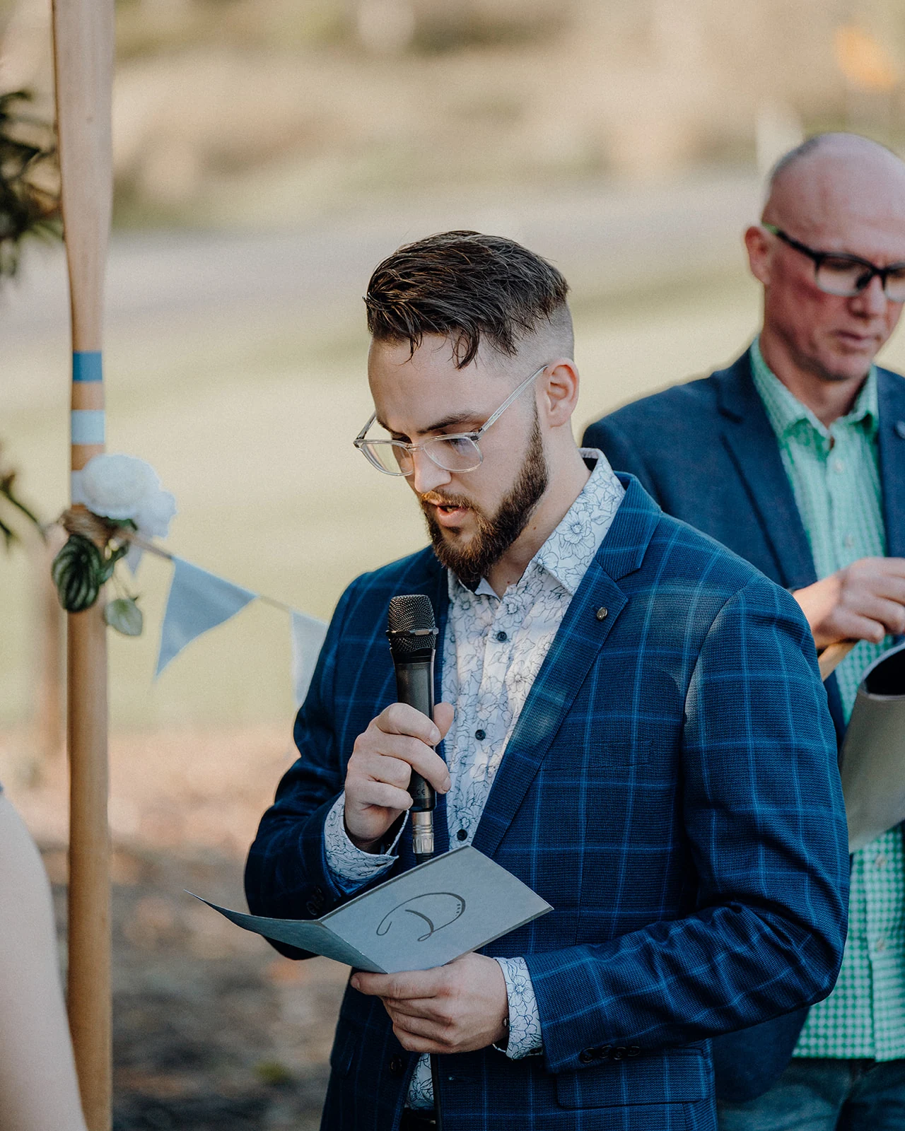 A groom reads his vows from a booklet at an outdoor ceremony.