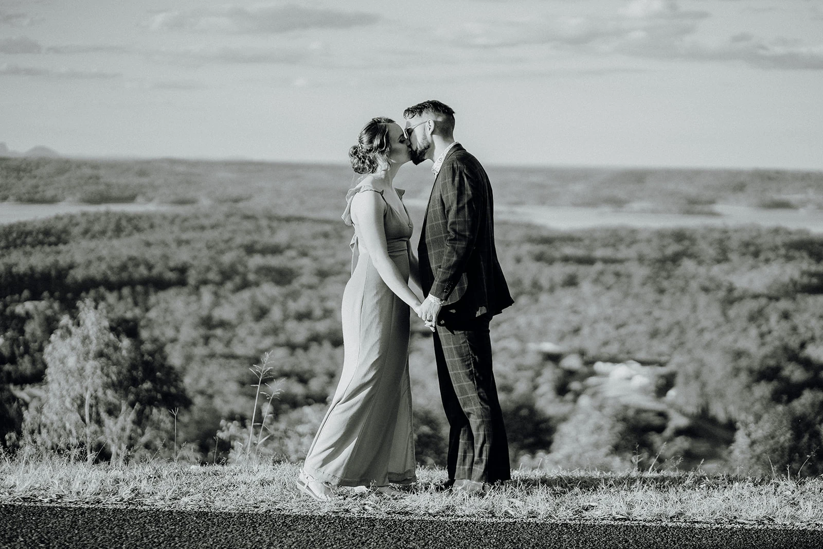 A couple share a kiss above a wooded valley, photographed in black and white.
