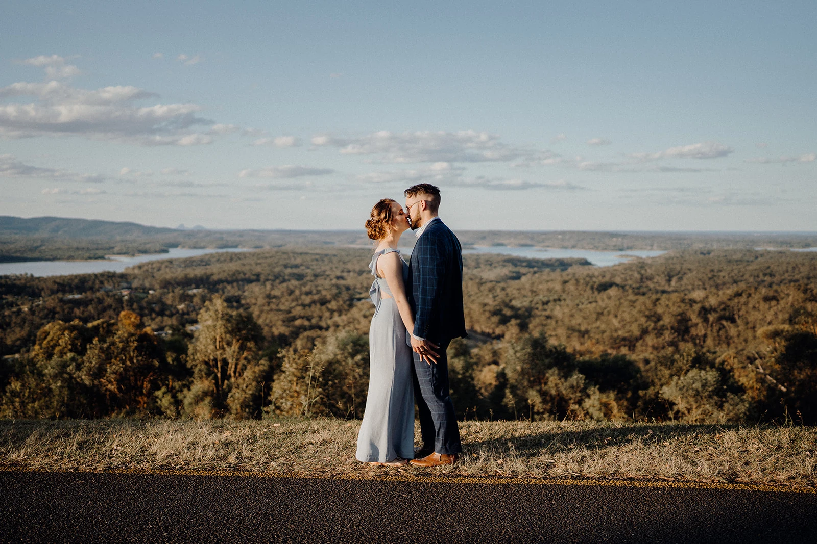 Newlyweds kiss above a forest and distant lake in warm afternoon light.