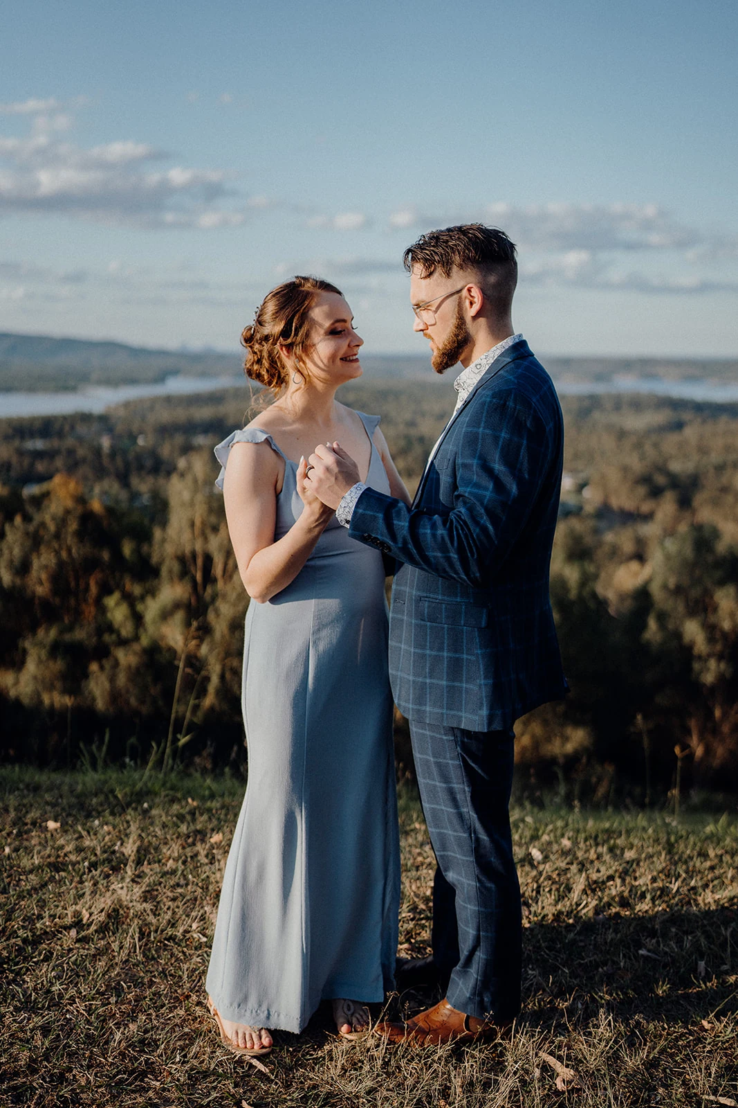 A laughing bride holds hands with her groom above a wooded valley.