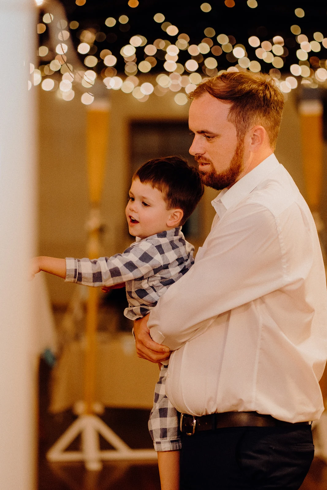 A man holds a young child beneath out-of-focus reception lights.