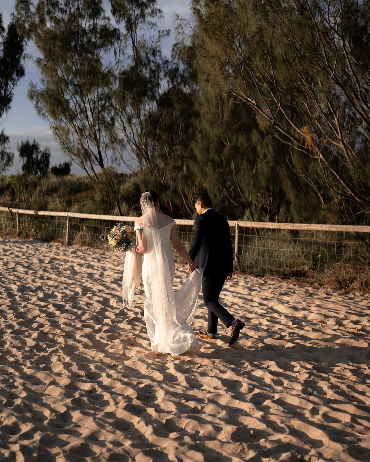 A bride and groom walk across sunlit sand beside coastal trees.