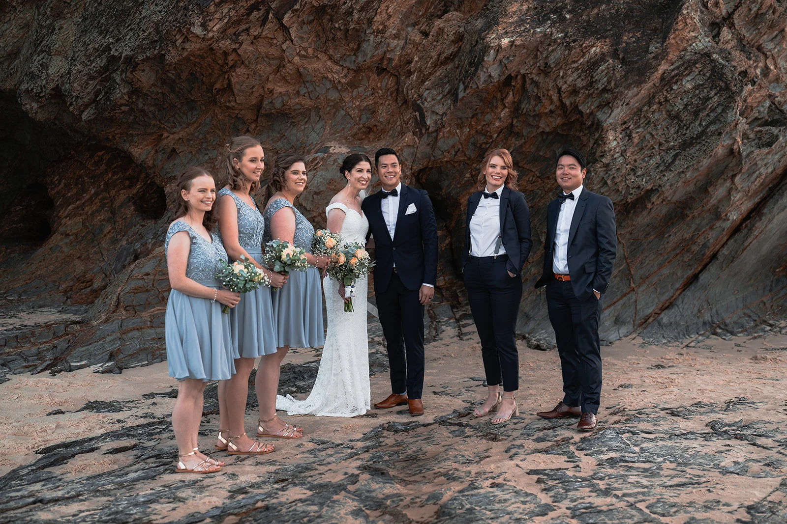 A wedding party gathers beneath a rocky coastal cliff.