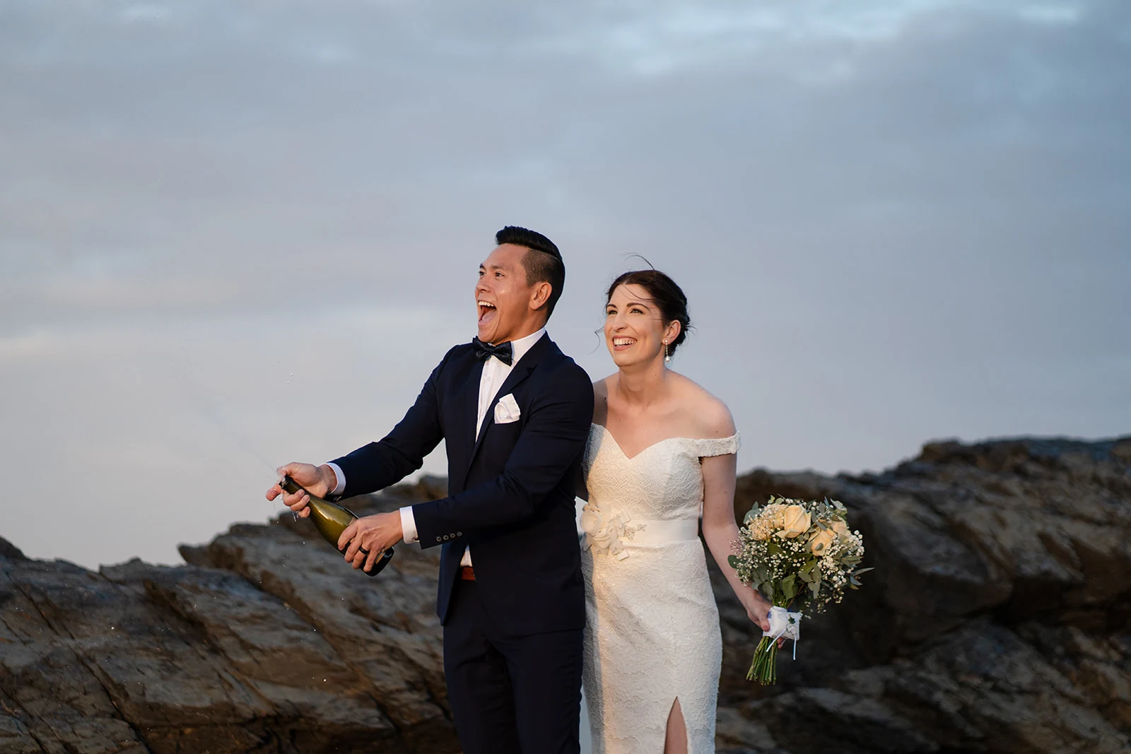 A groom opens a bottle of sparkling wine while his bride laughs beside him.