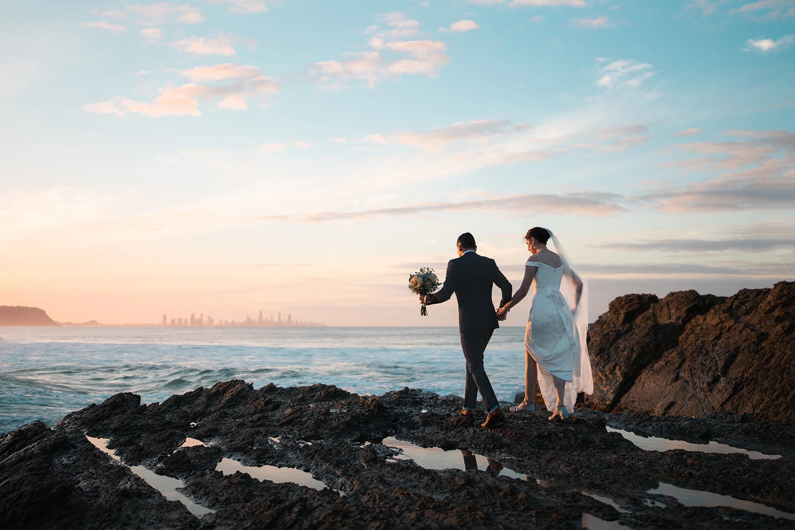 Newlyweds hold hands on coastal rocks beneath a pink and blue evening sky.