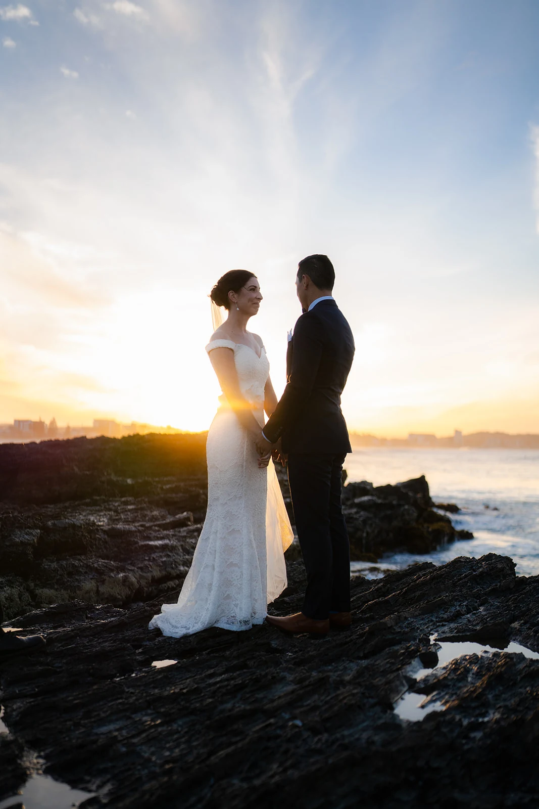 A low sun shines between a bride and groom standing beside the ocean.