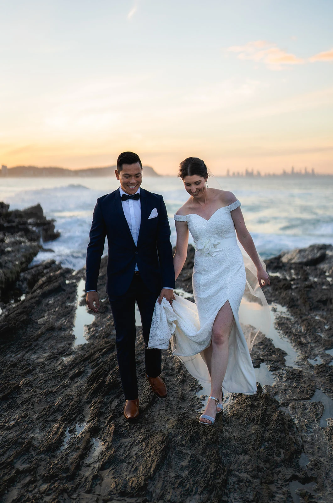 A couple walk hand in hand across coastal rocks at sunset.