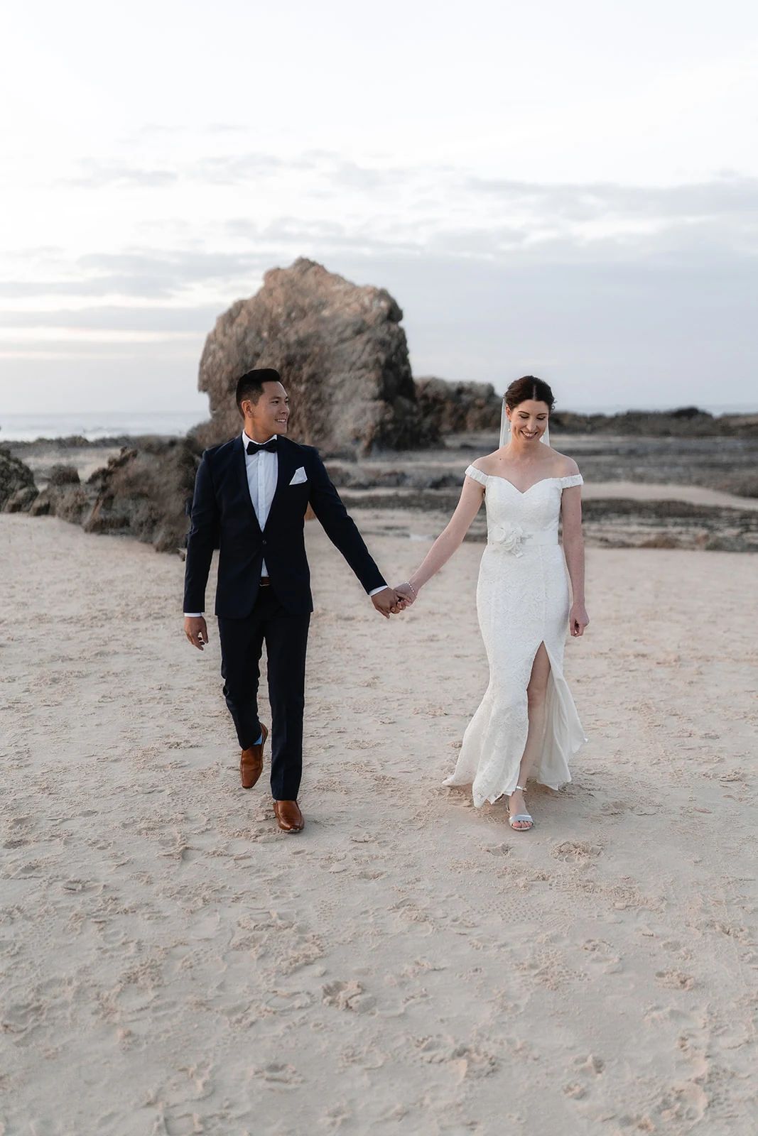 Newlyweds walk barefoot across the beach, holding hands.