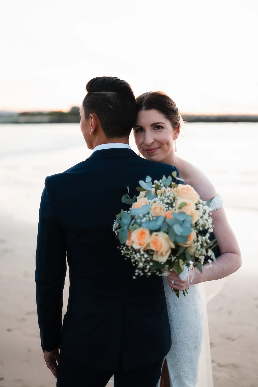 A bride looks over her groom's shoulder while holding a pastel bouquet.