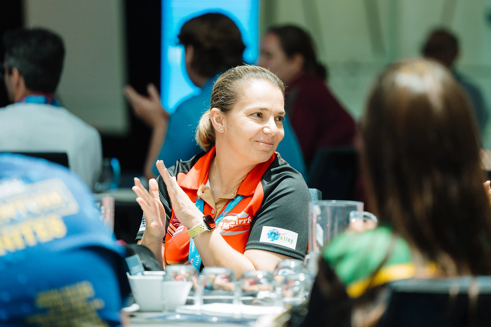 A woman applauds at a conference table, surrounded by other attendees.